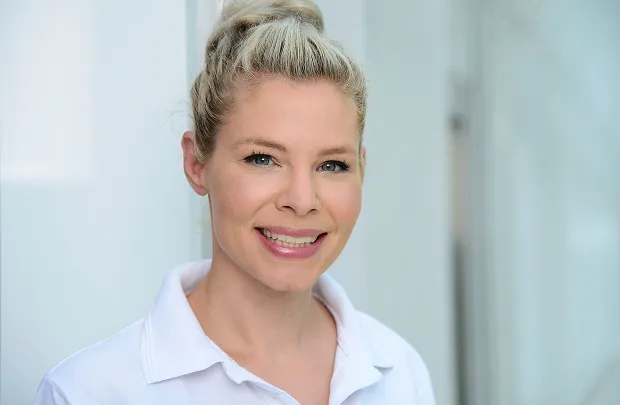 Porträt einer lächelnden Frau mit blonden, hochgesteckten Haaren vor hellem Hintergrund.
